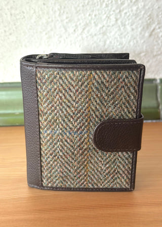 Sie sehen das Portemonnaie von vorne. Die Vorderseite ist schön mit Harris Tweed® ausgeführt. Es handelt sich um einen hellgrünen Fischgrät mit Musterkaro. Kombiniert wird der Harris Tweed® mit dunkelbraunem Leder. Eine Schließe aus Leder sichert das Portemonnaie.
