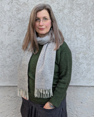 Im Bild zu sehen ist ein hellgrauer Schal mit Fischgrätmuster und cremefarbenen Fransen