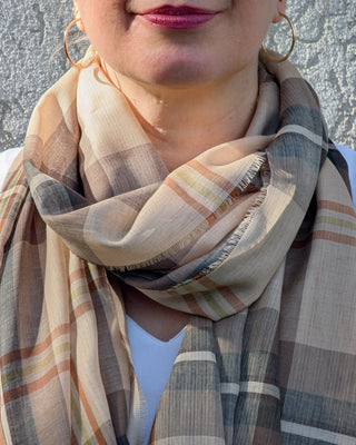 Zu sehen ist ein feiner karierter Merinoschal in den Farben beige, braun, weiß, schwarz, bronze in einer Schleife um einen Frauenhals gelegt.