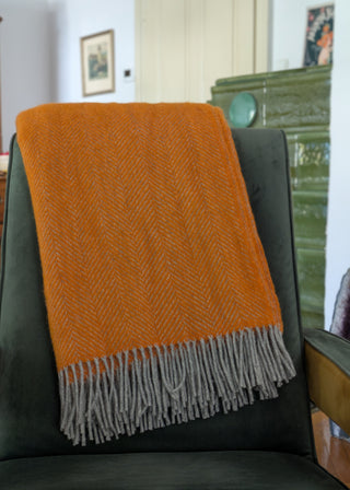 Zu sehen ist eine klassische Herringbone-Decke aus reiner Schurwolle in den Farben Orange & Grau. Sie ist über einen Lehnsessel gelegt. 