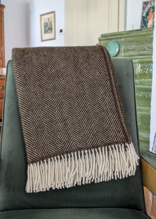 Zu sehen ist eine klassische Herringbone-Decke aus reiner Schurwolle in der Farbe Schoko. Sie ist über einen Lehnsessel gelegt. 